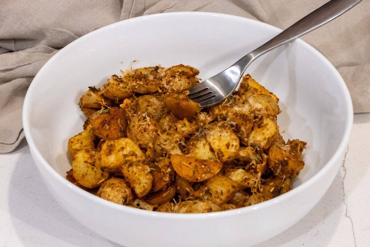A white bowl filled with seasoned roasted potatoes and herbs, with a fork resting on the side of the bowl.