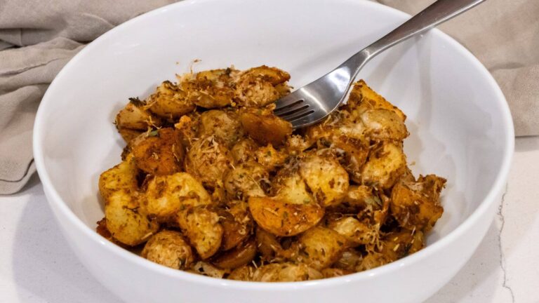 A white bowl filled with seasoned, roasted potato pieces, topped with herbs and grated cheese, with a fork resting on the edge.