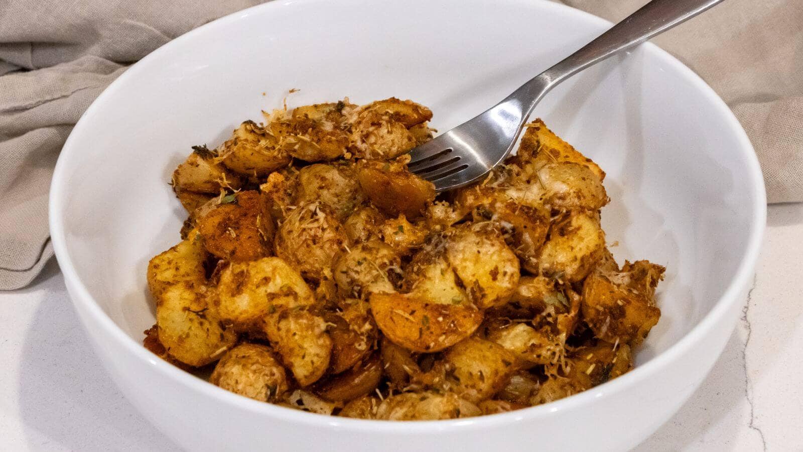 A white bowl filled with seasoned, roasted potato pieces, topped with herbs and grated cheese, with a fork resting on the edge.