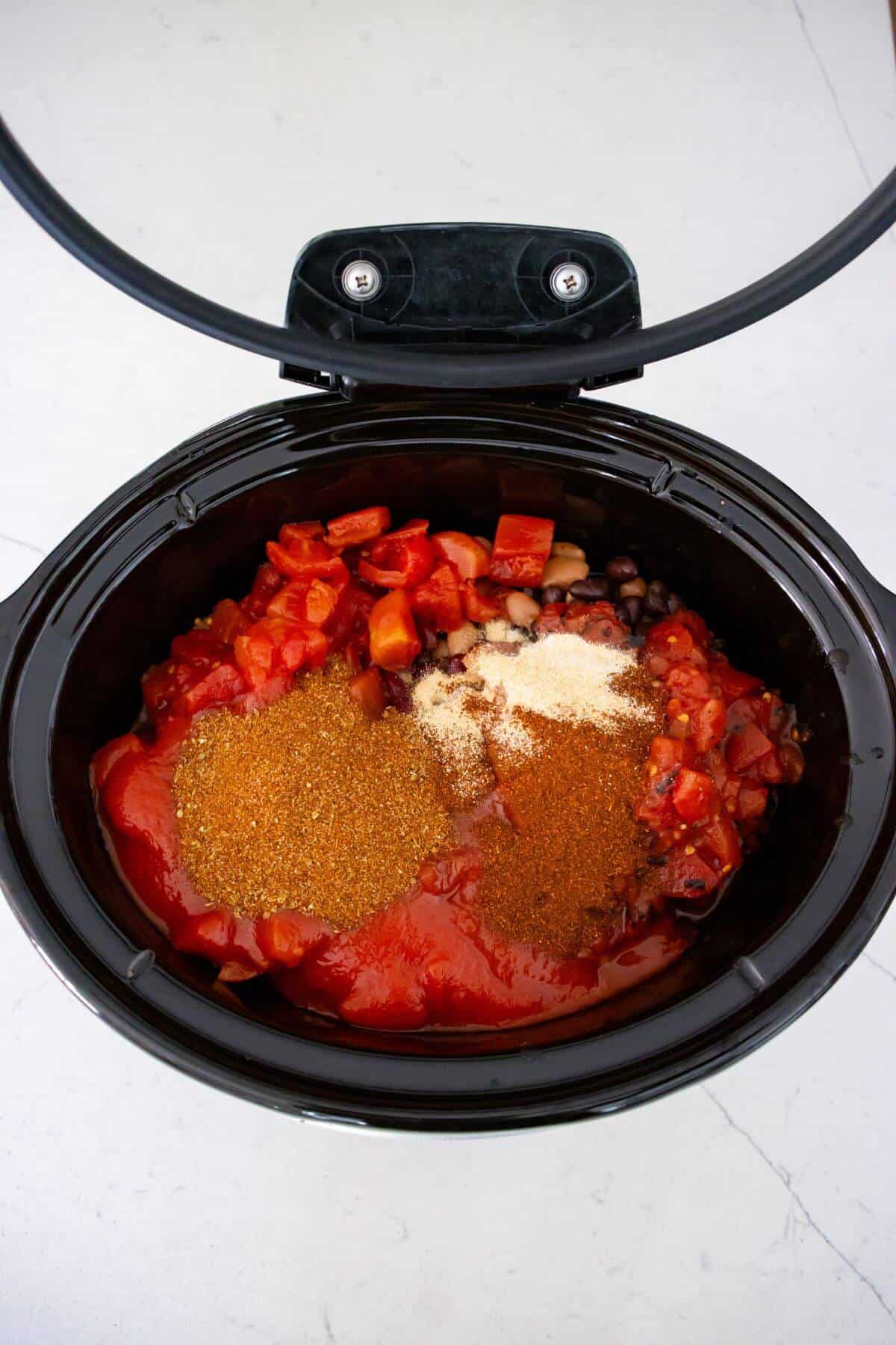 A slow cooker filled with diced tomatoes, beans, tomato sauce, and various spices, ready to be mixed and cooked.