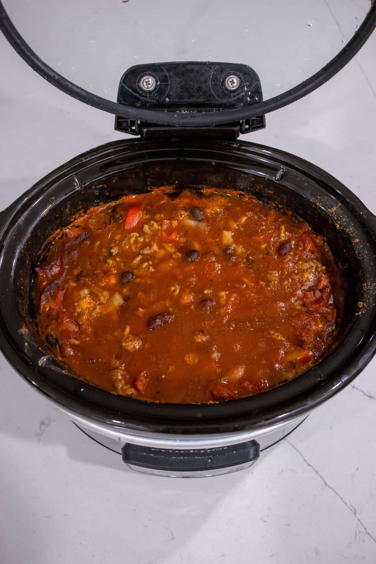 A slow cooker filled with a red, chunky stew containing beans, tomatoes, and pieces of vegetables, sitting on a light-colored countertop.