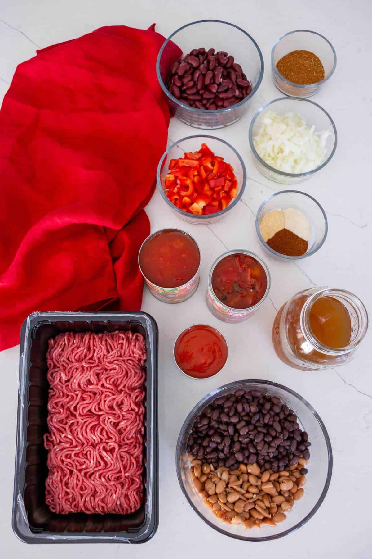 A variety of chili ingredients on a countertop, including ground beef, beans, canned tomatoes, bell pepper, onion, spices, and broth in bowls and containers.