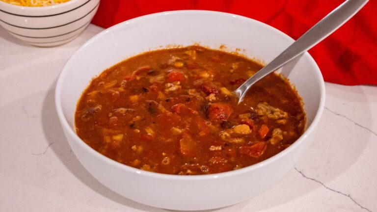 A white bowl filled with chunky chili or stew, containing ground meat, beans, and tomatoes, with a metal spoon inside.