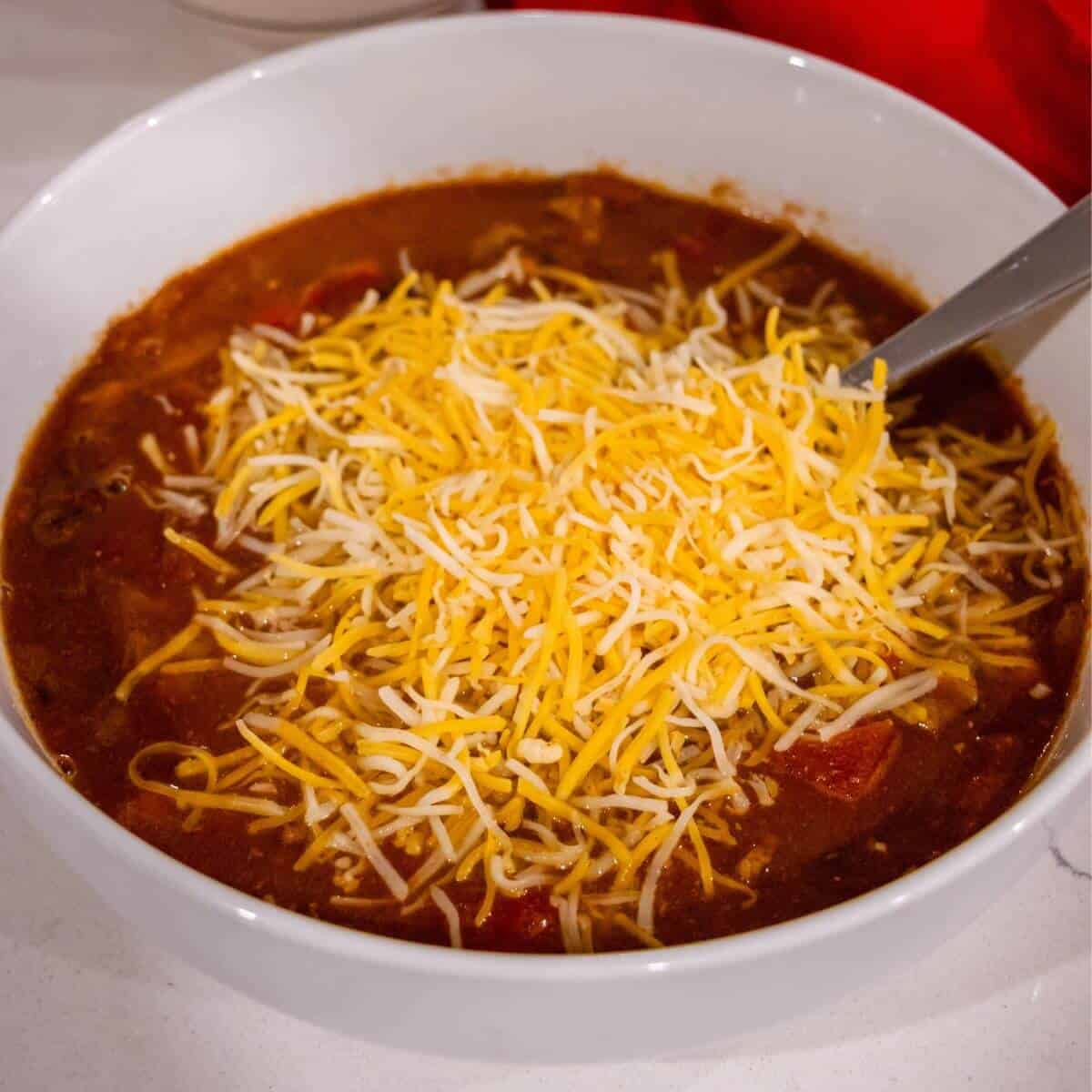 A white bowl filled with chili topped with a generous amount of shredded cheddar and Monterey Jack cheese, with a spoon resting inside.