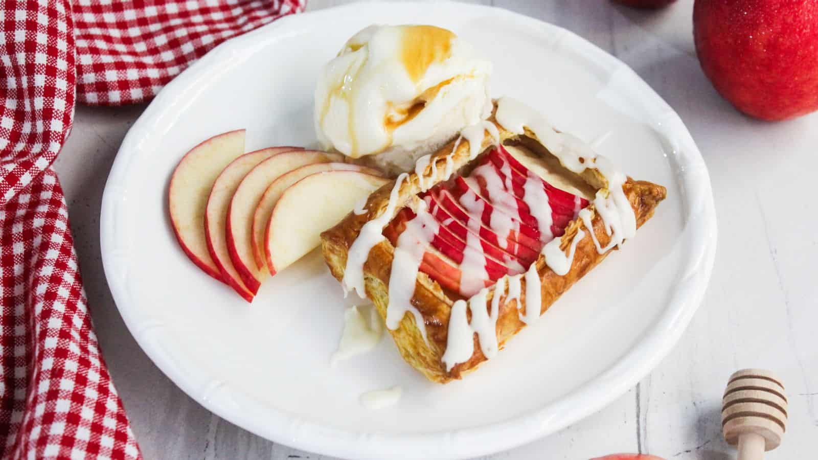 A plate with a slice of apple pastry drizzled with white icing, a scoop of vanilla ice cream with honey, and thin apple slices. A red checkered cloth is beside the plate.