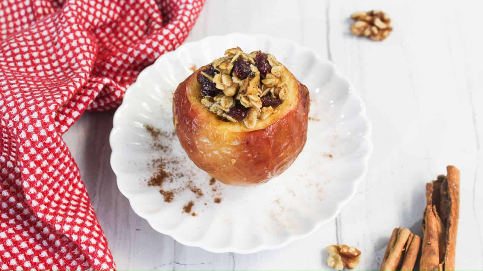 A baked apple filled with walnuts and cranberries on a white plate, sprinkled with cinnamon, with a red patterned cloth and cinnamon sticks nearby.
