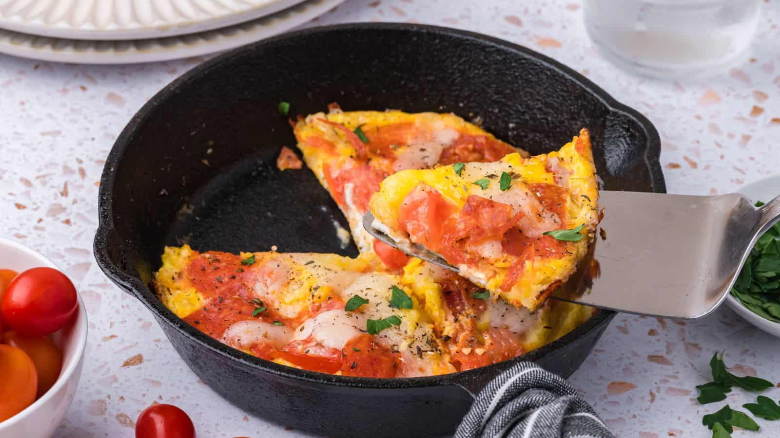 A slice of tomato and cheese frittata is being lifted from a cast iron skillet with a spatula.