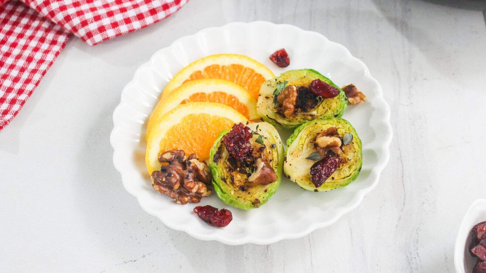 A white plate with three orange slices and three Brussels sprout halves topped with walnuts and dried cranberries, on a white surface with a red checkered cloth.