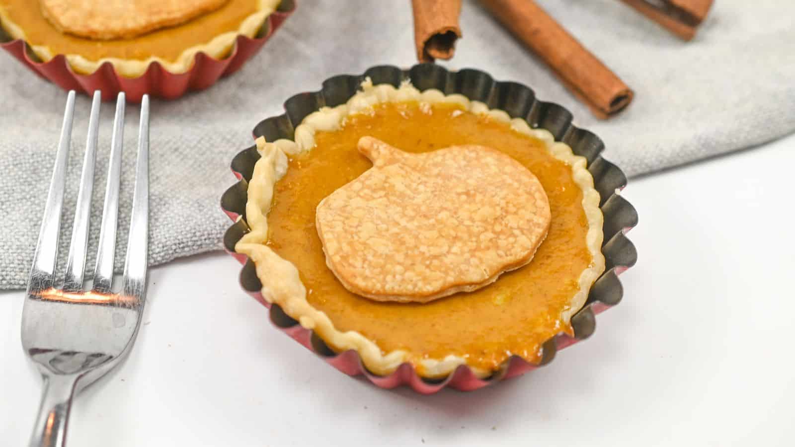 Mini pumpkin pie in a tart pan topped with a pumpkin-shaped piece of pastry, with a fork and cinnamon sticks nearby on a gray napkin.