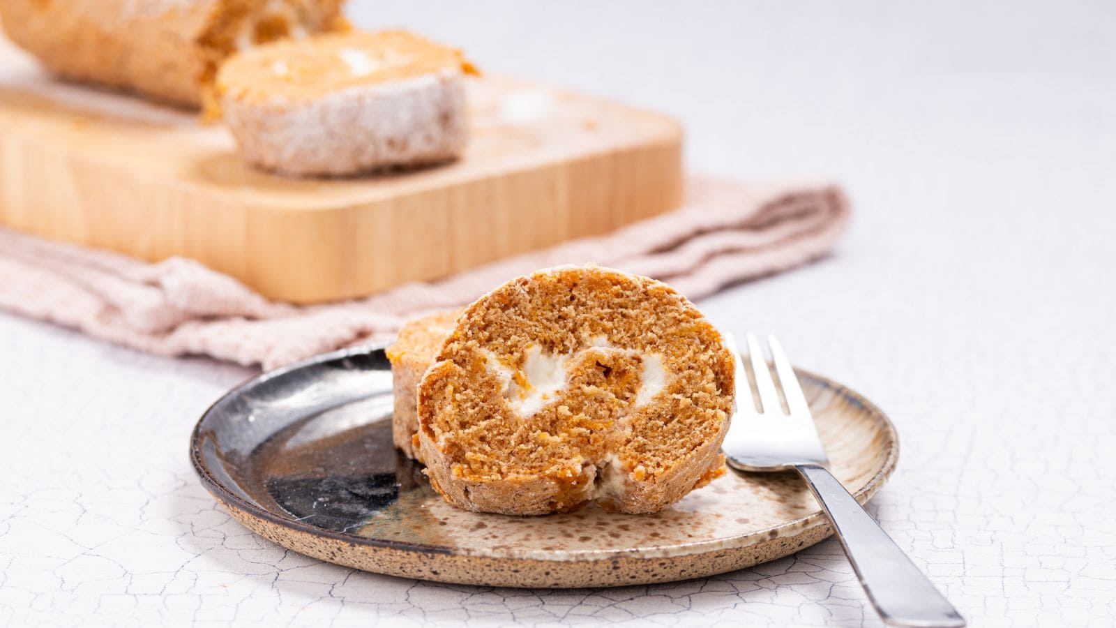 A slice of pumpkin roll cake with cream cheese filling is served on a plate with a fork; more slices are visible on a cutting board in the background.