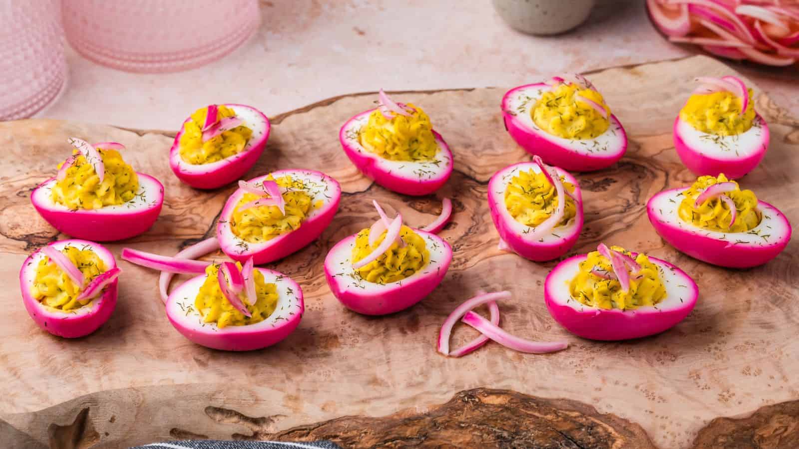 Deviled eggs with bright pink-colored whites, yellow yolk filling, and garnished with dill and pickled onions, arranged on a wooden serving board.