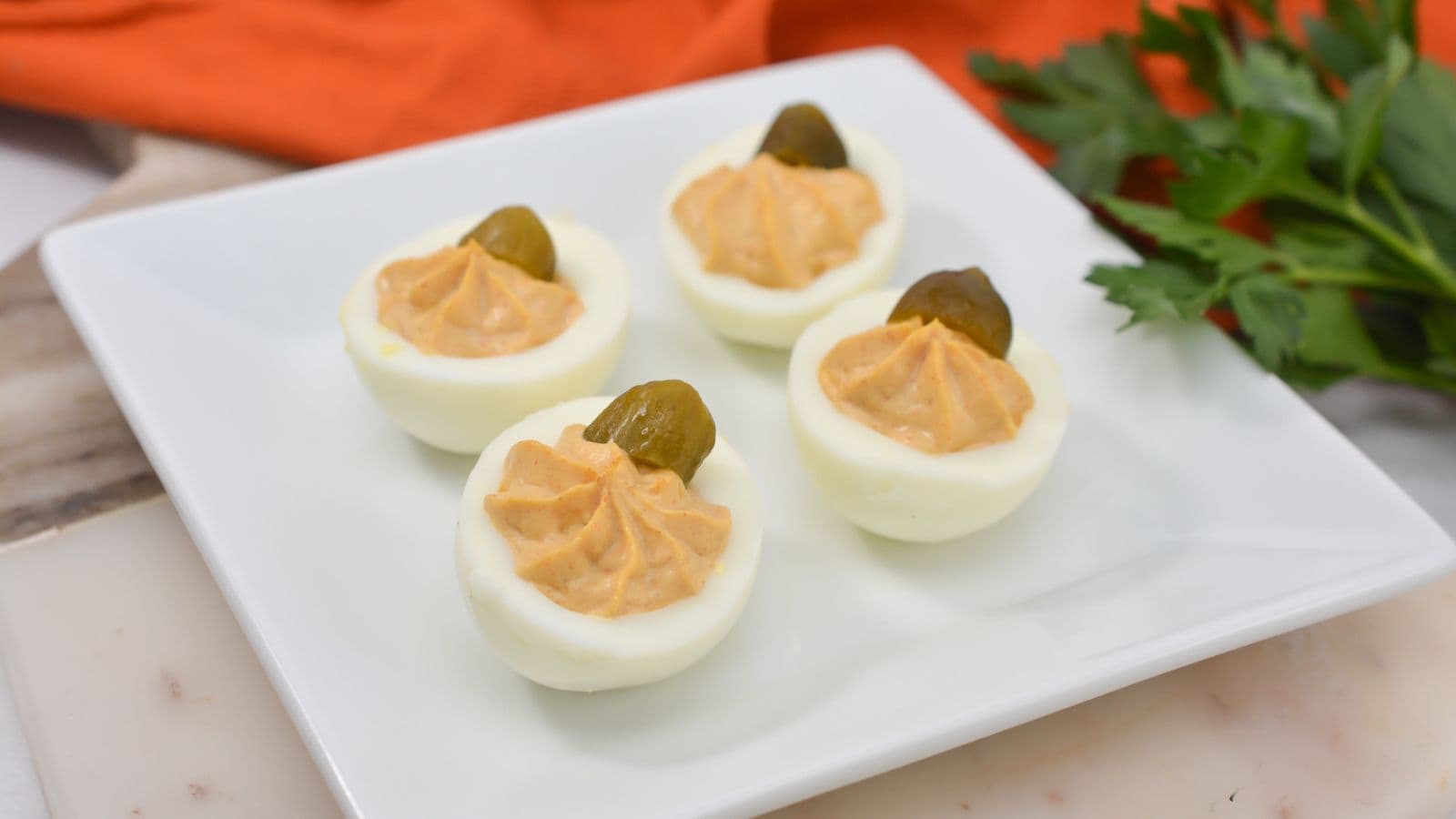 Four deviled eggs topped with a small pickle slice each, arranged on a white square plate with parsley and an orange napkin in the background.