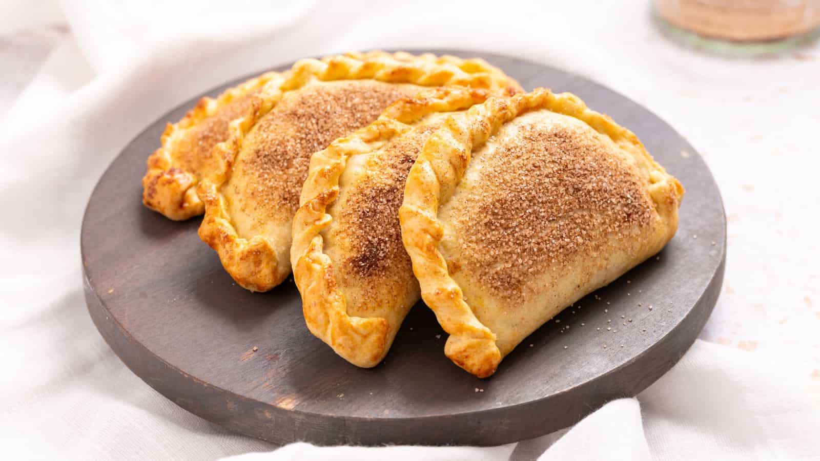 Three baked hand pies with golden, crimped edges and a dusting of cinnamon sugar are arranged on a dark round plate.