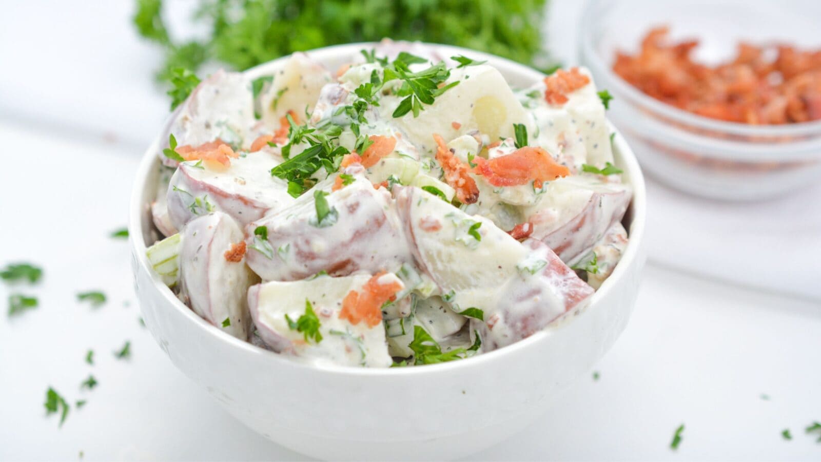 A bowl of creamy potato salad topped with chopped parsley and bacon pieces, with extra bacon and herbs visible in the background.