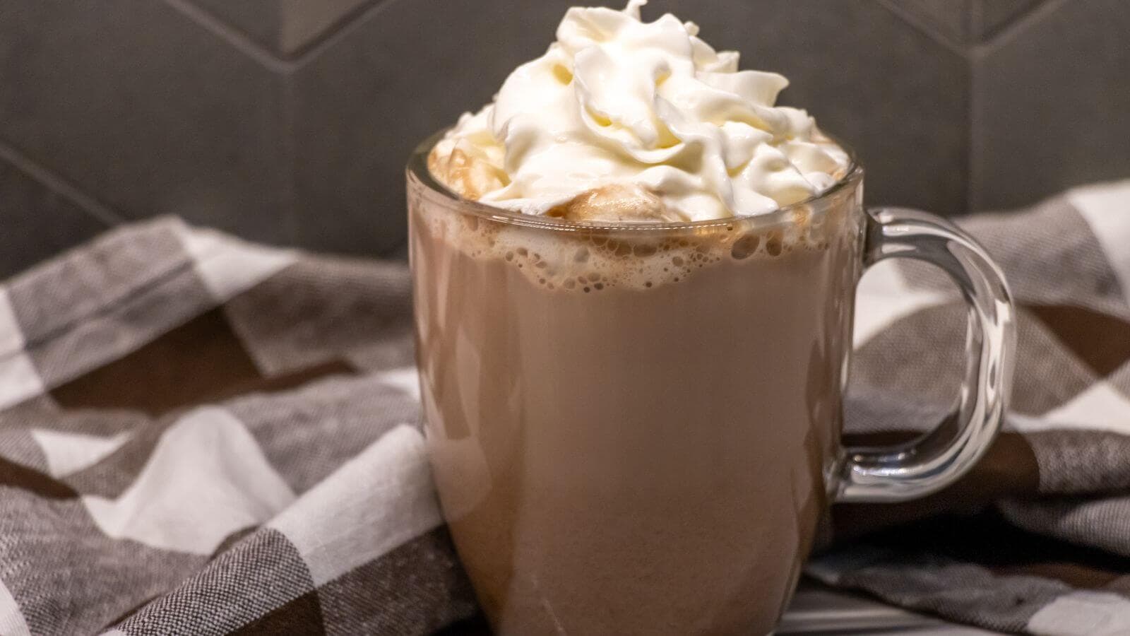 A clear glass mug filled with hot chocolate topped with whipped cream, placed in front of a brown and white checkered cloth.