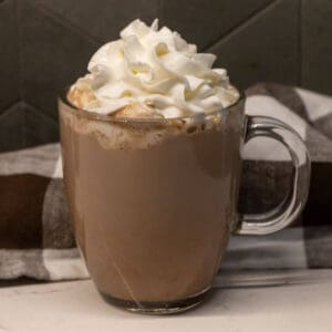 A clear glass mug filled with hot chocolate, topped with whipped cream, sits on a white surface in front of a striped cloth.