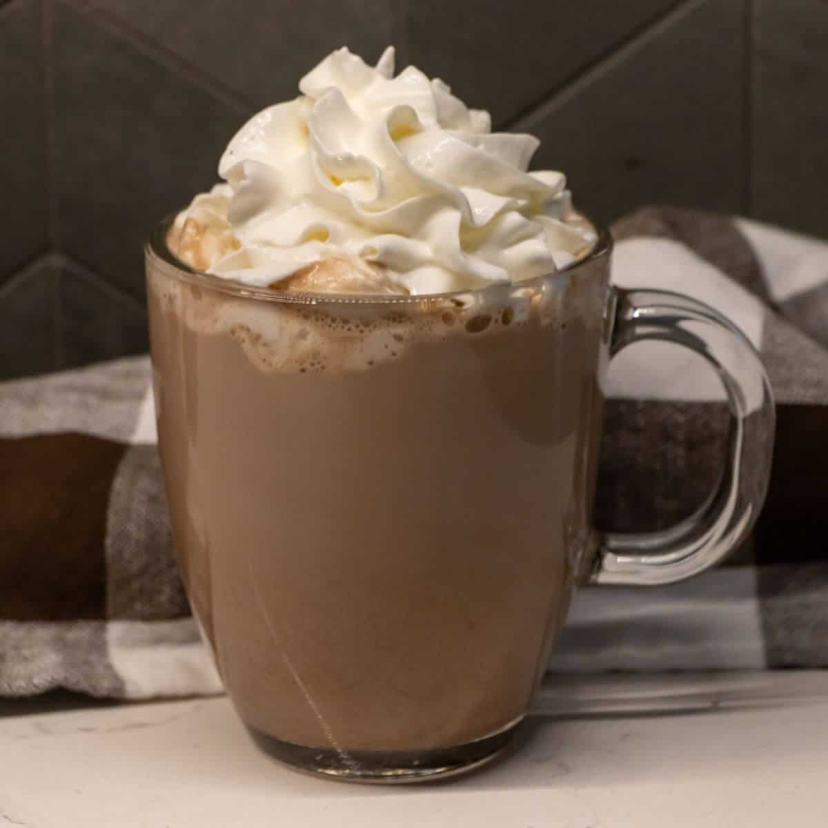 A clear glass mug filled with hot chocolate, topped with whipped cream, sits on a white surface in front of a striped cloth.