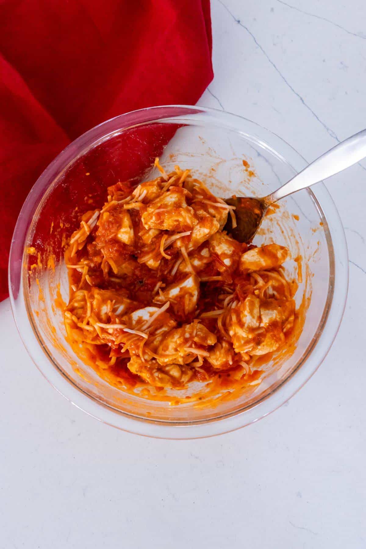 A glass bowl filled with shredded chicken mixed with red sauce and cheese, with a spoon inside, sits on a white surface next to a red cloth.