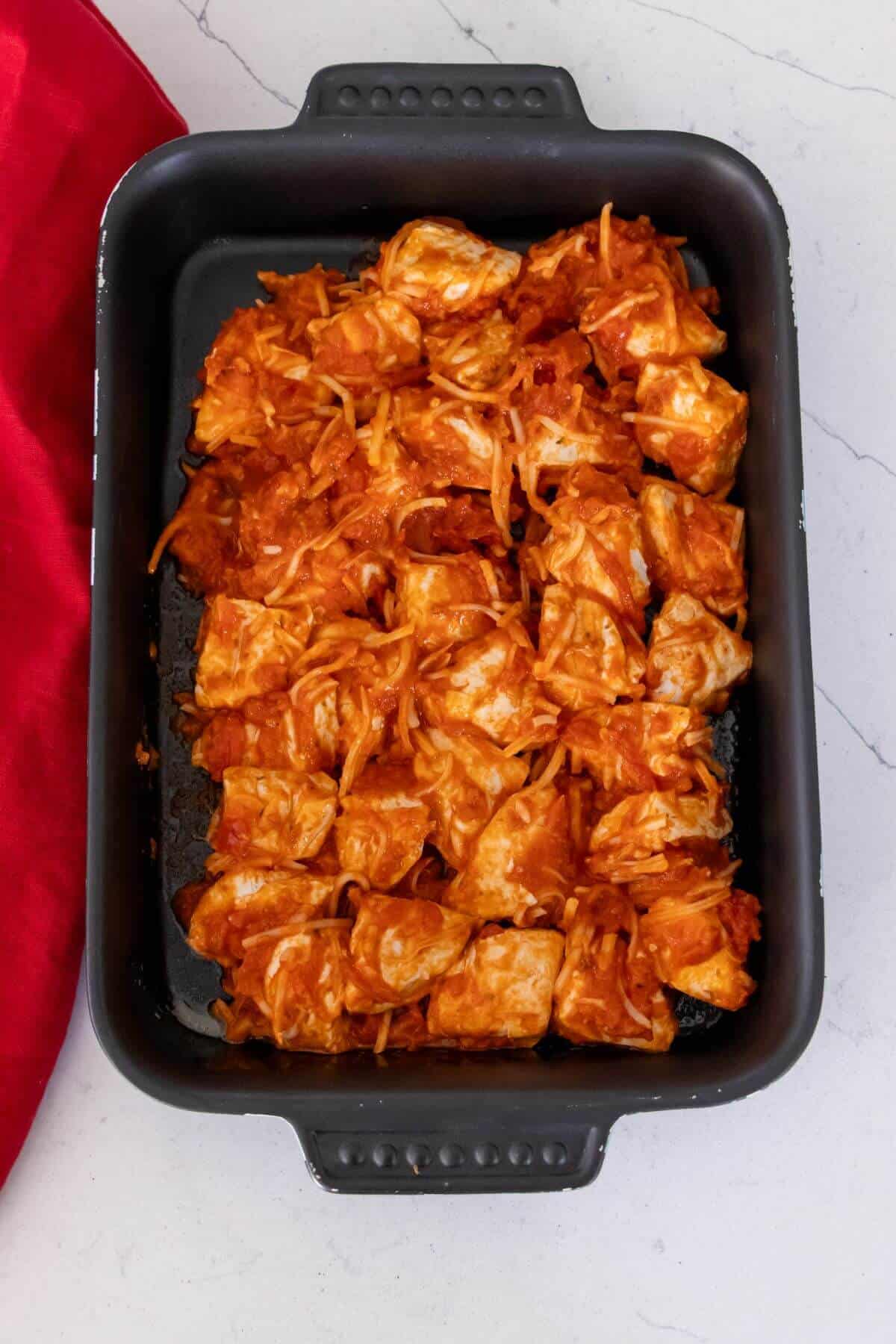 A black baking dish filled with cubed pieces of lasagna roll-ups mixed with tomato sauce and cheese, placed on a white counter next to a red cloth.