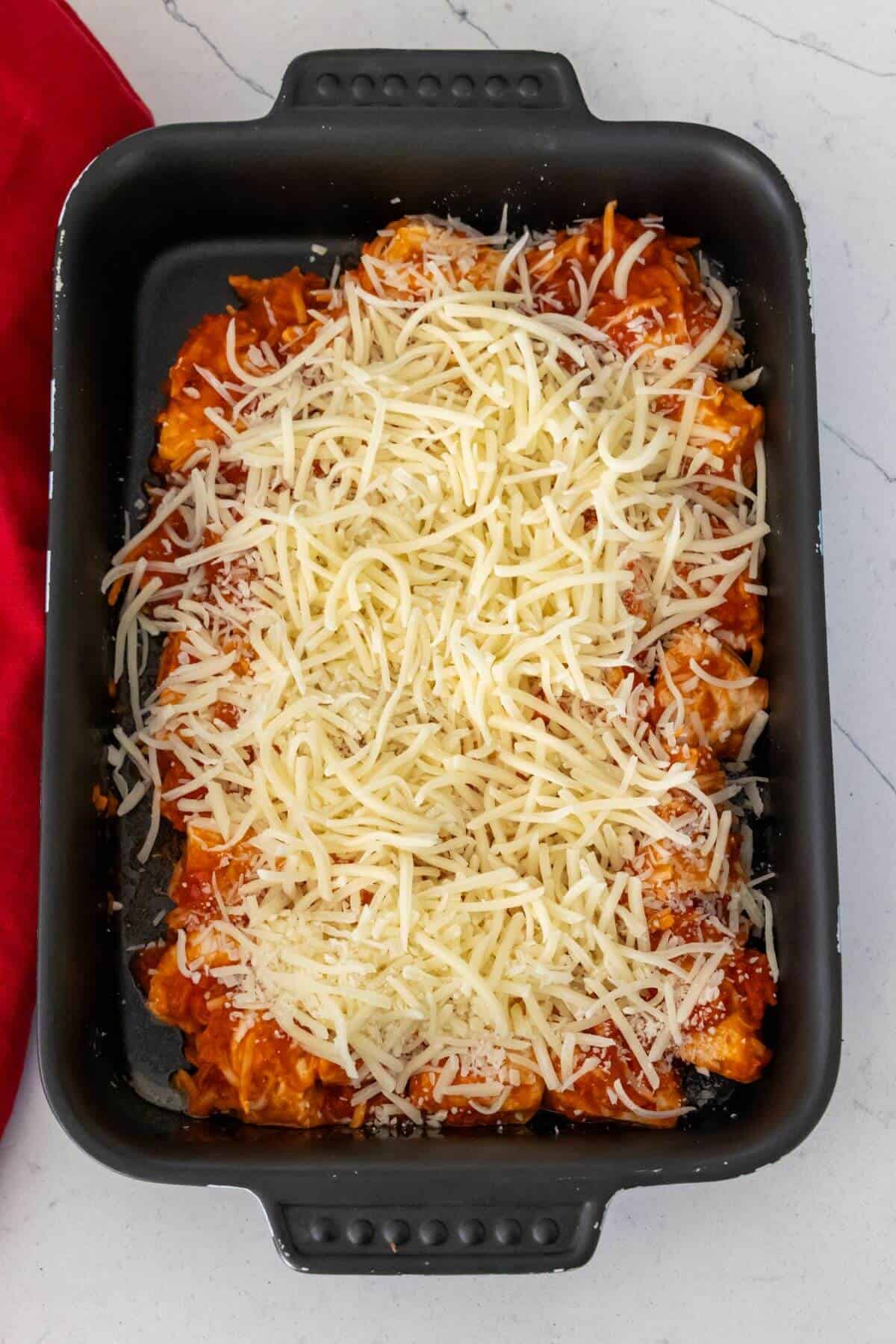 A black baking dish filled with uncooked pasta topped with tomato sauce and a layer of shredded cheese on a white countertop.