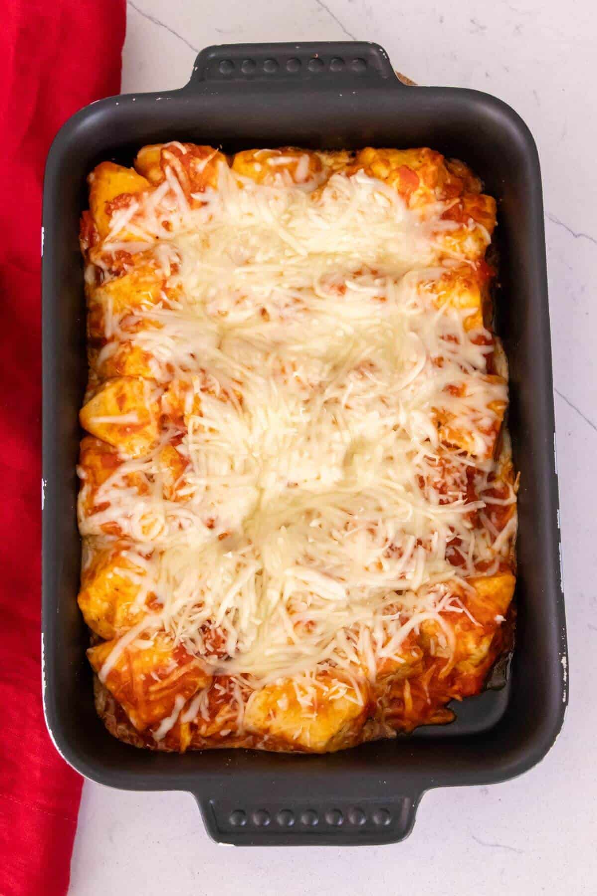 A black baking dish filled with enchiladas covered in tomato sauce and melted shredded cheese, placed on a light surface next to a red cloth.