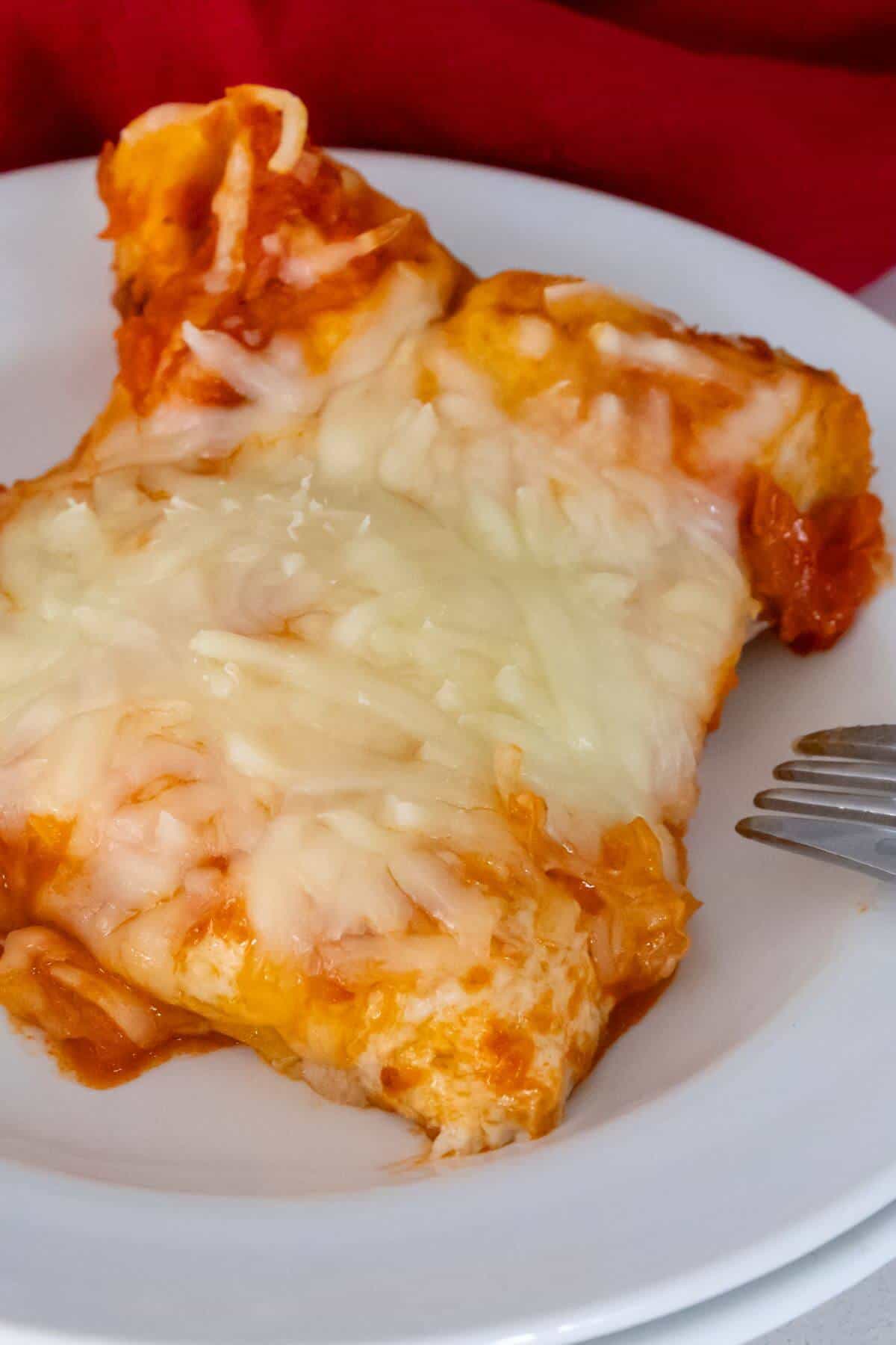 A slice of cheesy lasagna with melted cheese on top, served on a white plate with a fork beside it.