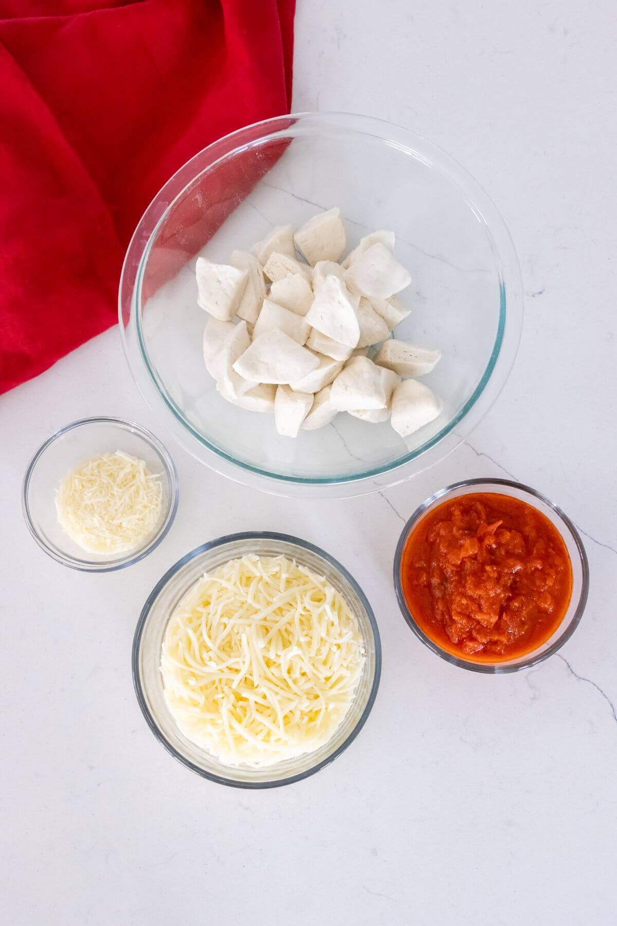 Four bowls on a white surface containing biscuit dough pieces, tomato sauce, shredded mozzarella cheese, and grated parmesan cheese; a red cloth is in the corner.