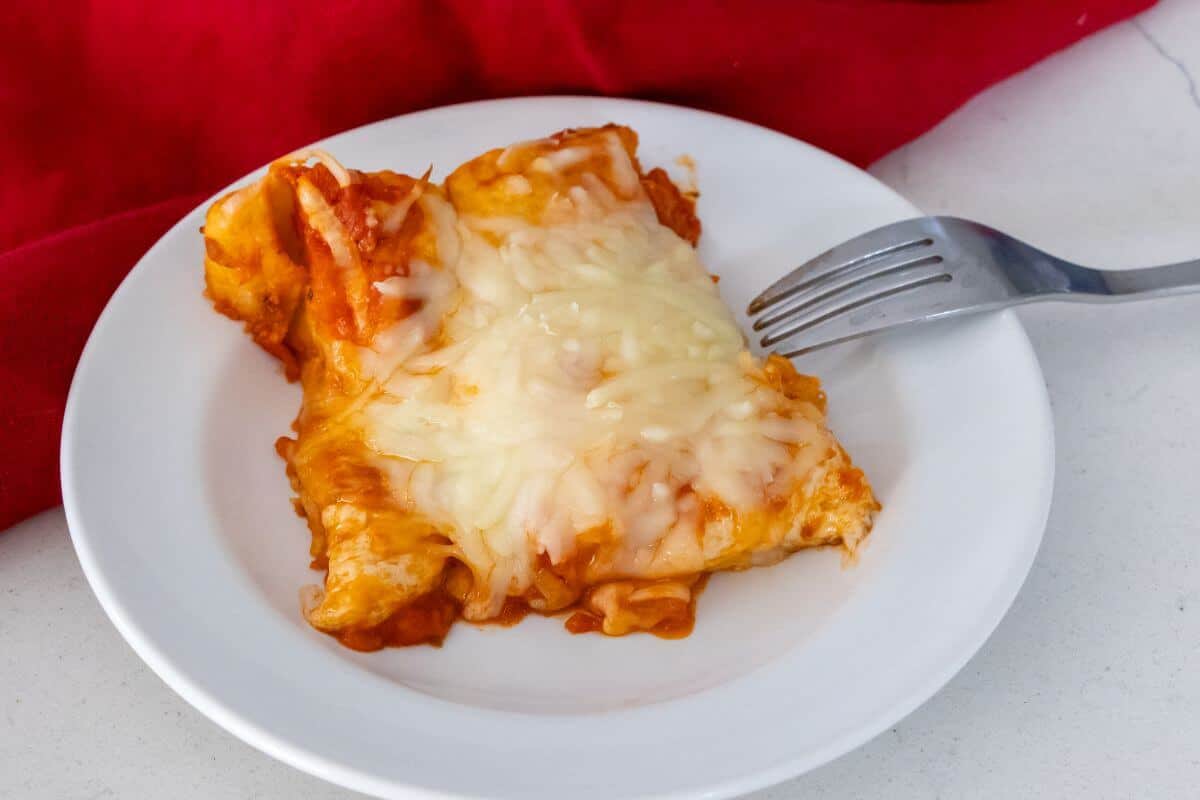 A slice of cheesy enchilada with melted cheese on top is served on a white plate with a fork beside it.