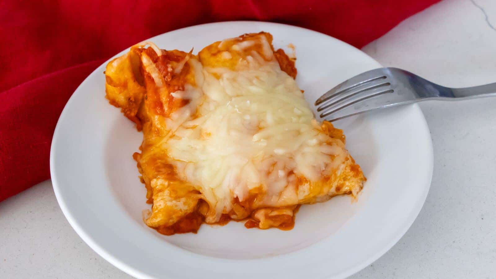 A serving of cheesy enchiladas with melted cheese on a white plate, next to a fork and a red cloth.