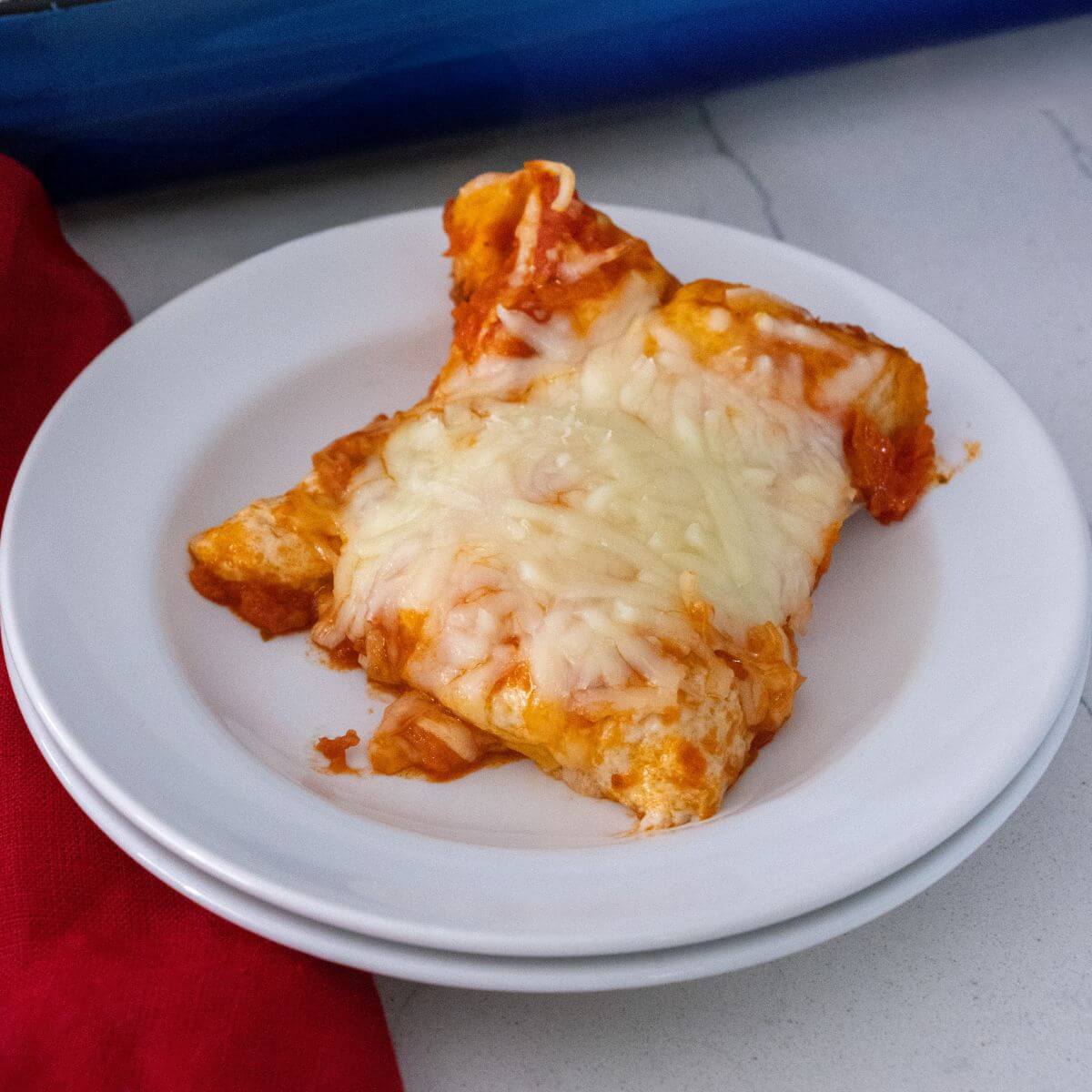 A serving of cheesy baked lasagna on a white plate, set on a white surface with a red cloth partially visible.
