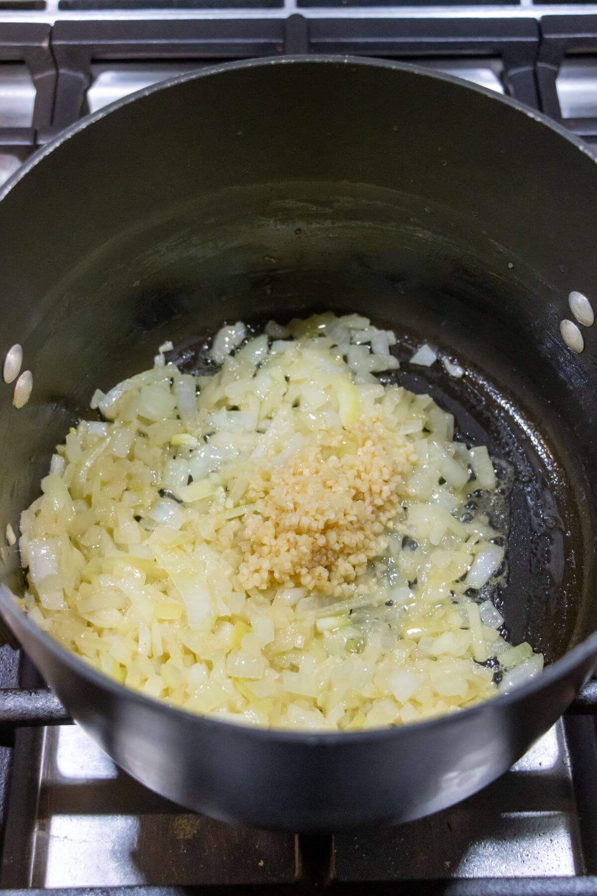 Chopped onions and minced garlic are being sautéed in a black pot on a stovetop.