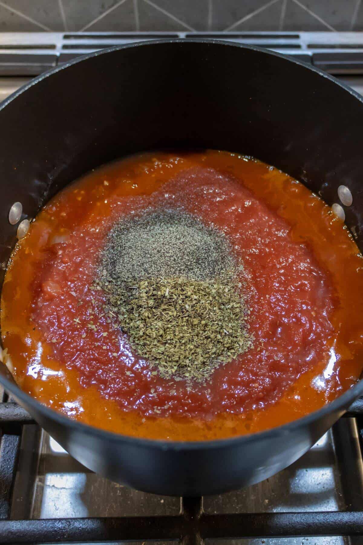 A pot of tomato sauce is heating on a stovetop with dried herbs and black pepper sprinkled on top, ready to be mixed in.