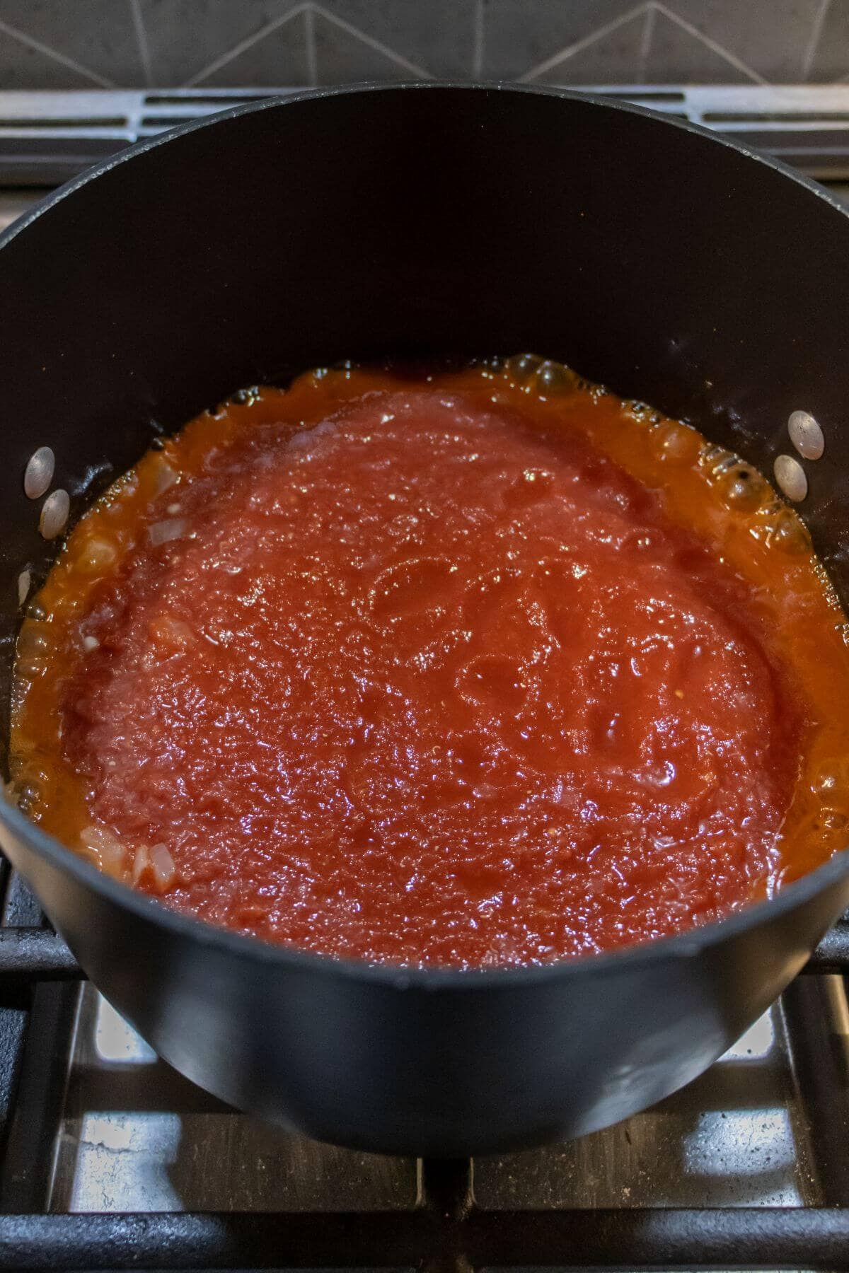 A pot of tomato sauce simmers on a stovetop, with visible diced onions around the edges of the sauce.