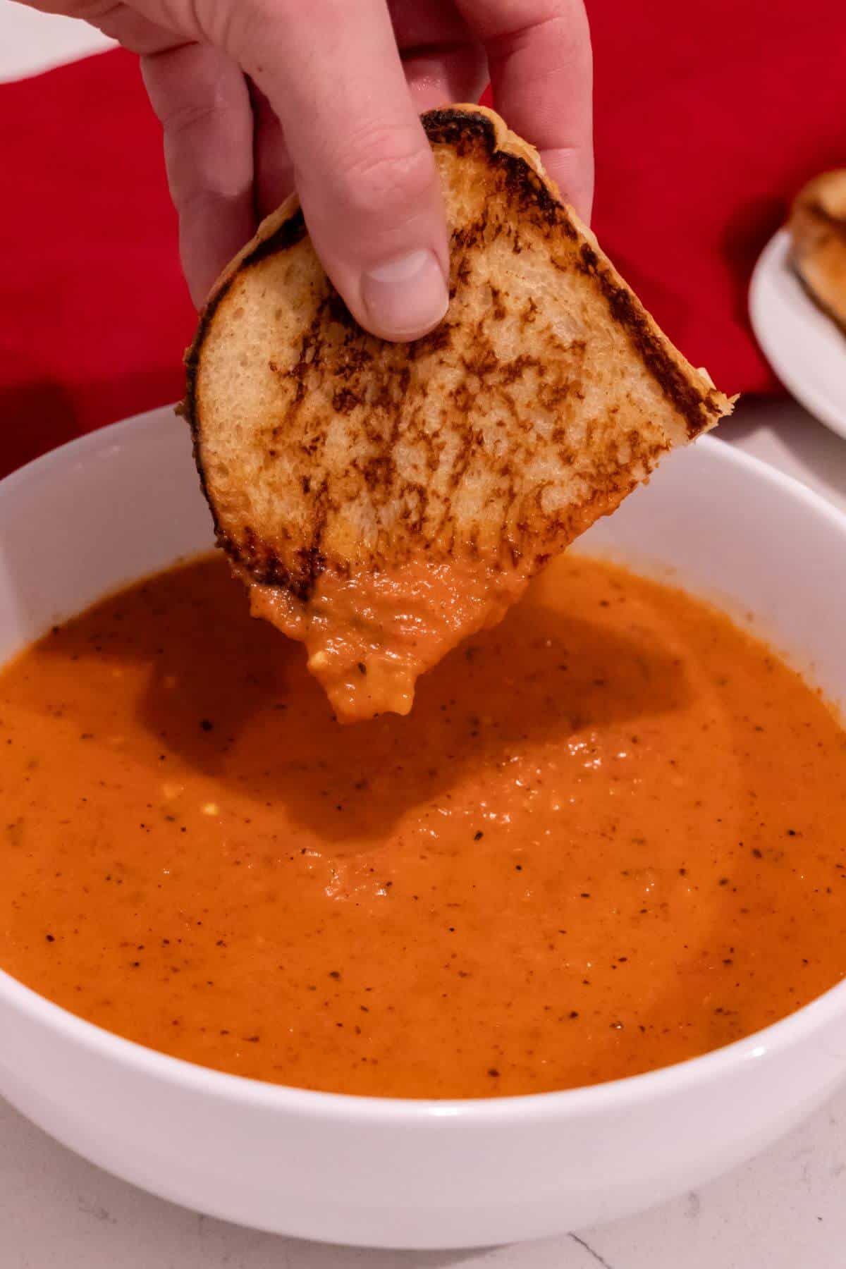 A hand dipping a grilled cheese sandwich into a bowl of tomato soup, with a red cloth in the background.