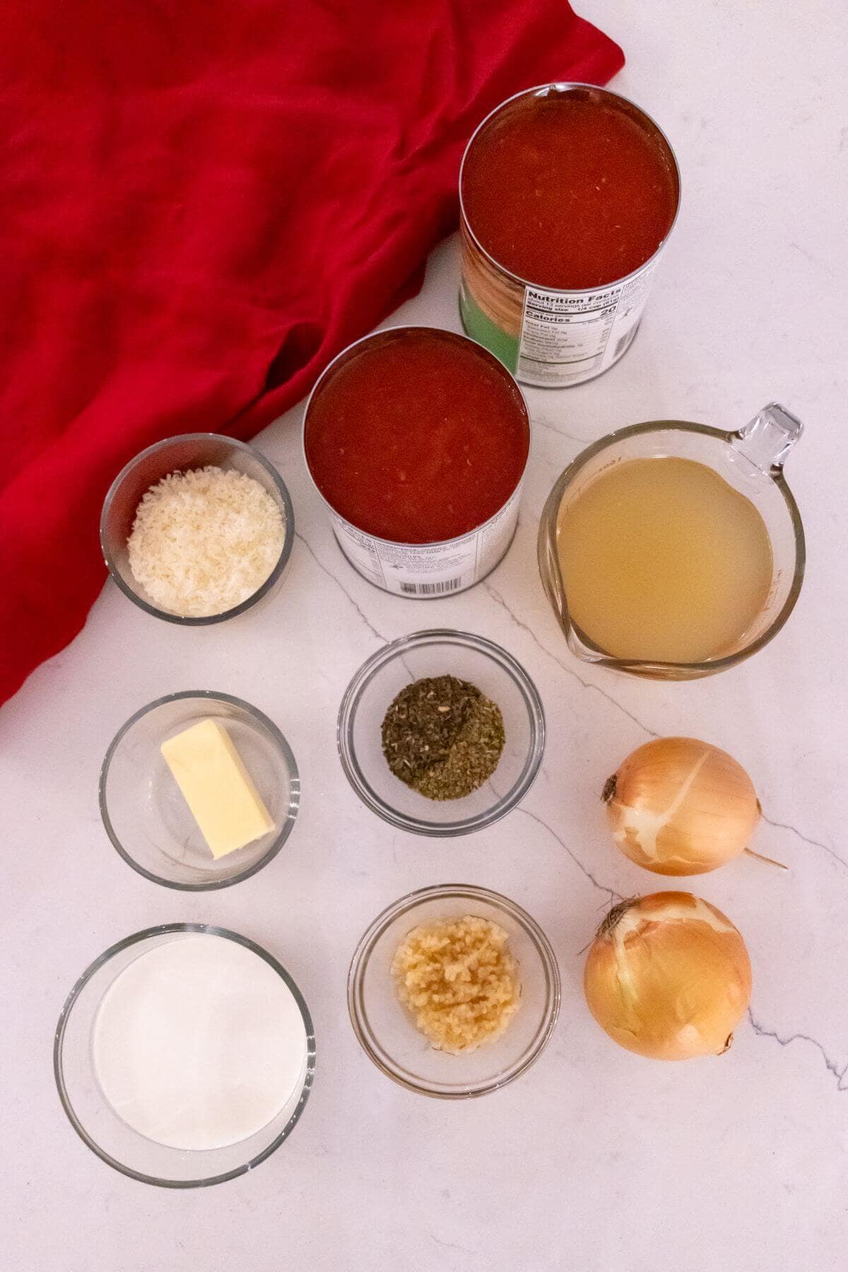 Ingredients arranged on a white surface: canned tomatoes, vegetable broth, onions, garlic, dried herbs, butter, heavy cream, grated cheese, and a red cloth.