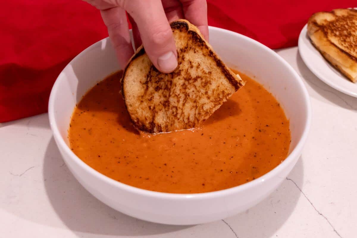 A hand dips a grilled cheese sandwich into a bowl of tomato soup on a white countertop.