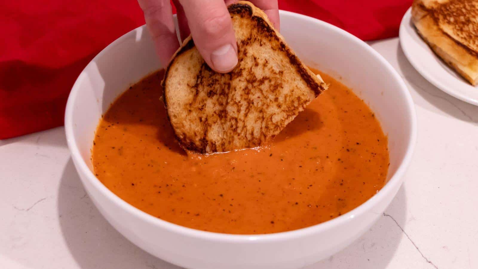 A hand dips a piece of grilled cheese sandwich into a bowl of tomato soup on a white countertop.