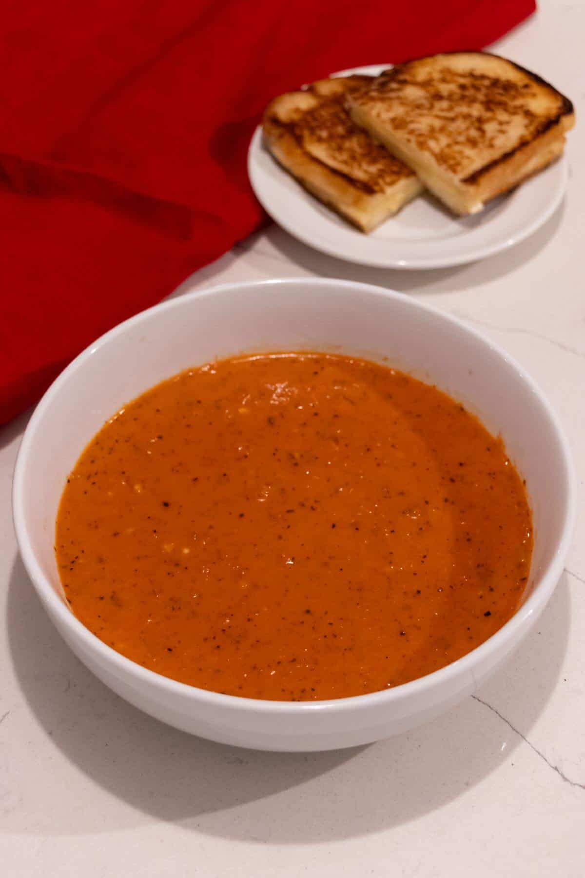 A white bowl of tomato soup sits on a countertop with a plate of grilled cheese sandwiches in the background.