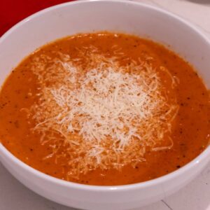 A bowl of creamy tomato soup topped with shredded cheese, served in a white bowl on a light surface.