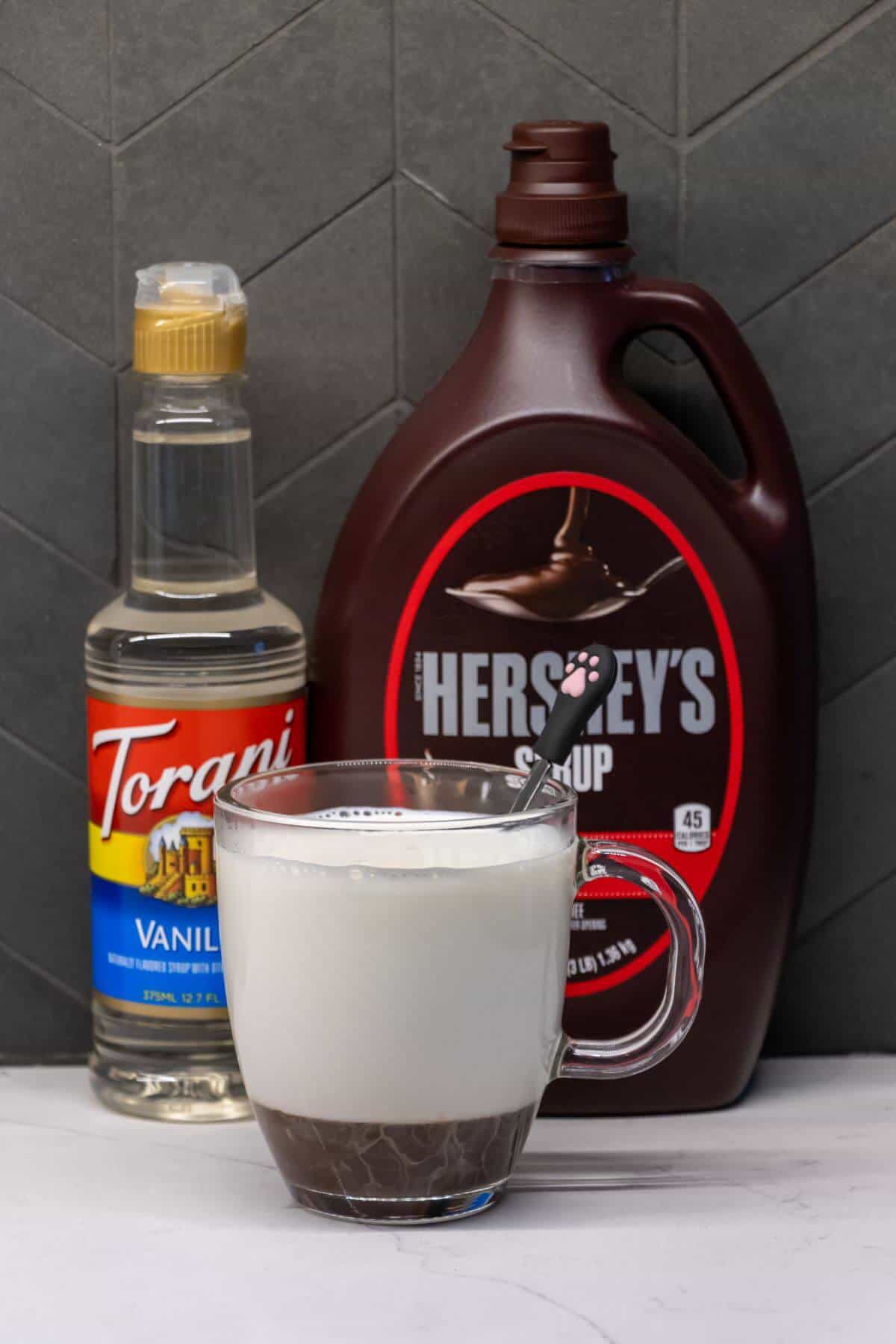 A glass mug with milk and chocolate syrup sits in front of a bottle of vanilla syrup and a bottle of Hershey’s chocolate syrup, with a spoon in the mug.