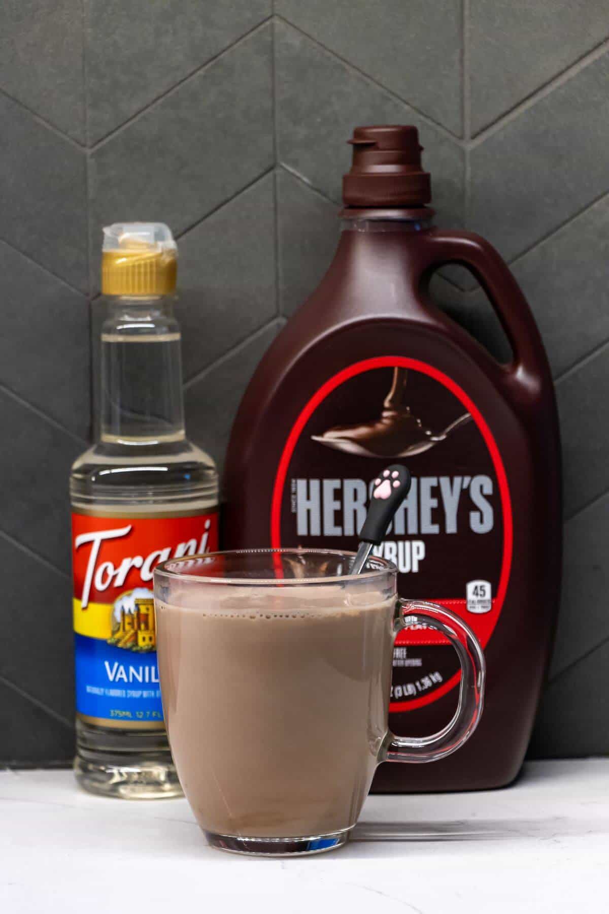 A glass mug of chocolate milk with a spoon inside, placed in front of bottles of vanilla syrup and Hershey's chocolate syrup on a gray tiled background.