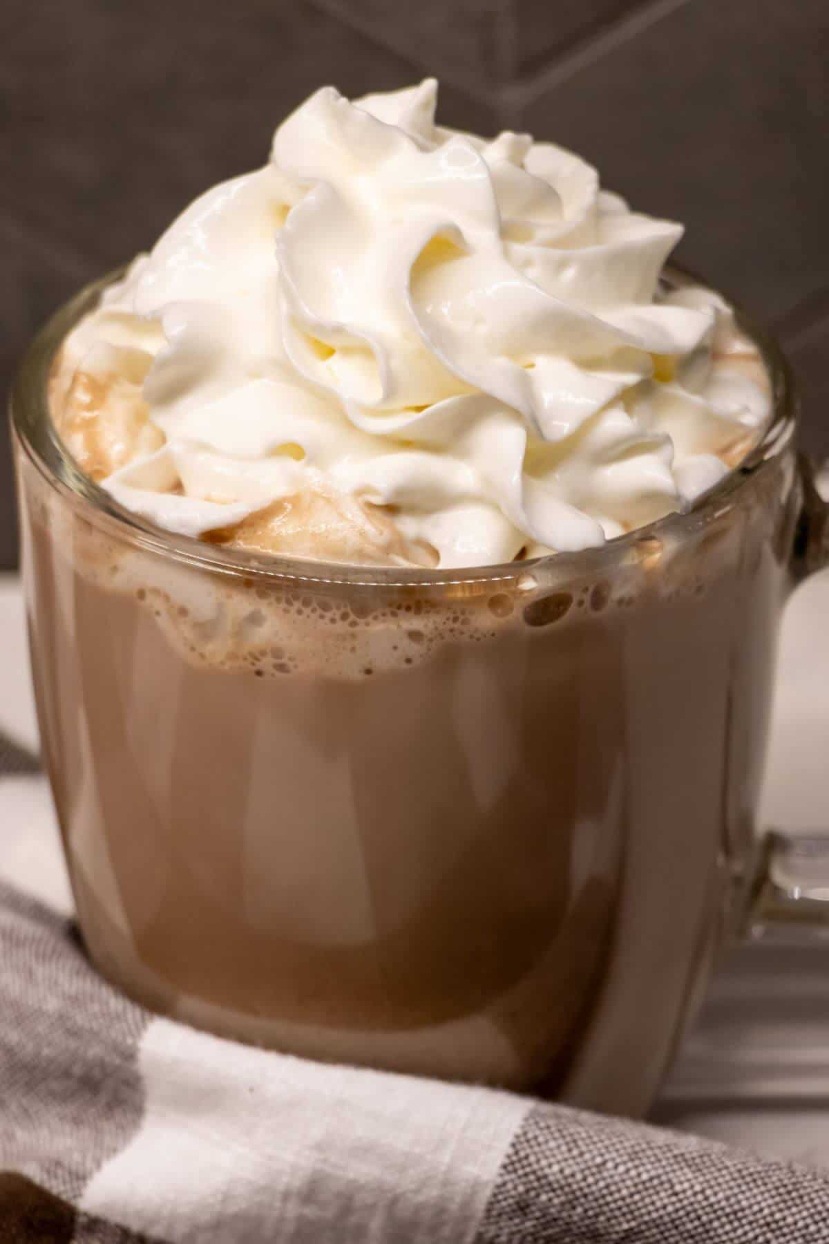 A glass mug filled with hot chocolate topped with a generous swirl of whipped cream, with a striped cloth nearby.