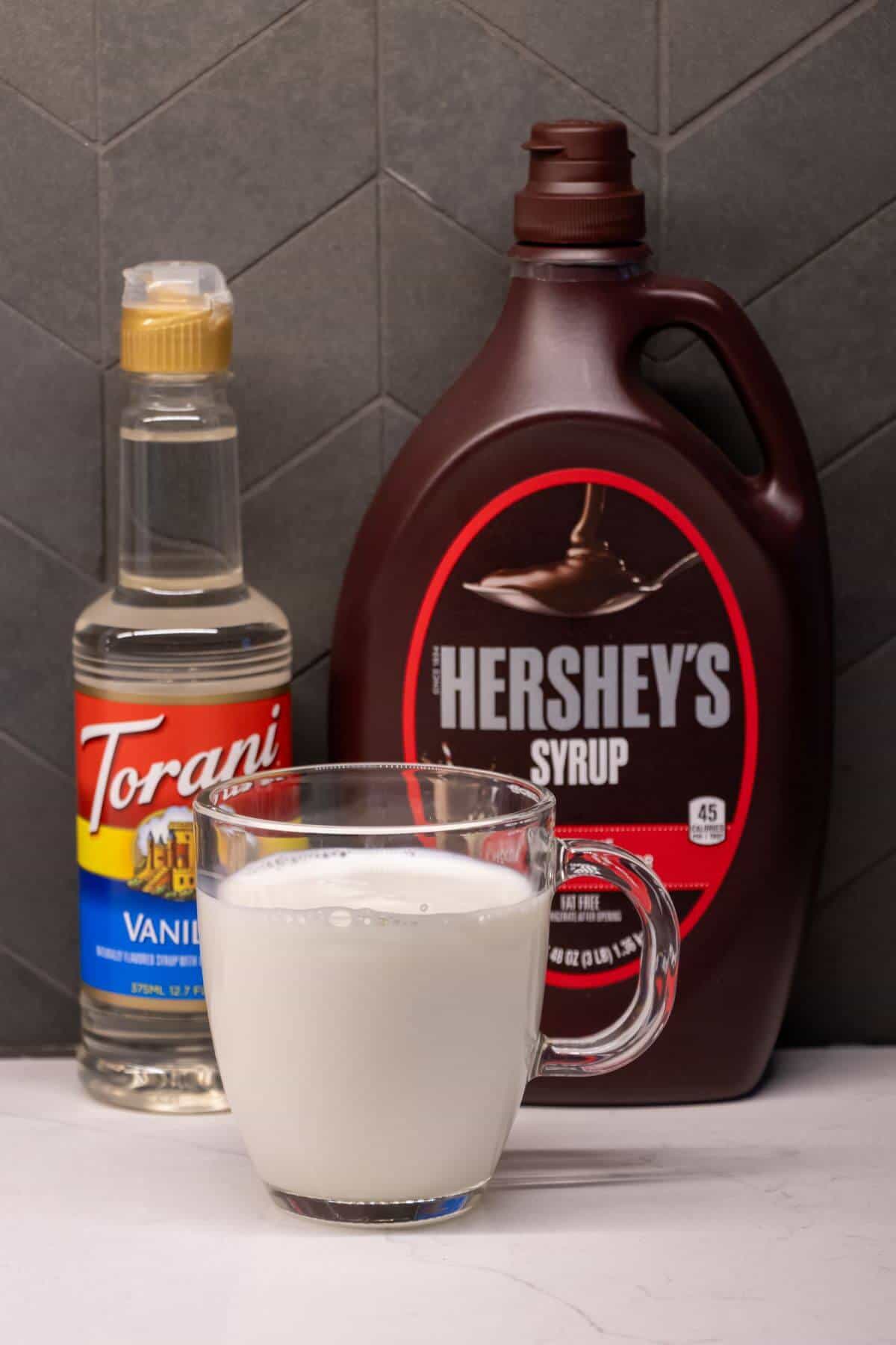 A glass mug of milk sits in front of a bottle of Torani vanilla syrup and a large bottle of Hershey’s chocolate syrup on a gray tiled surface.