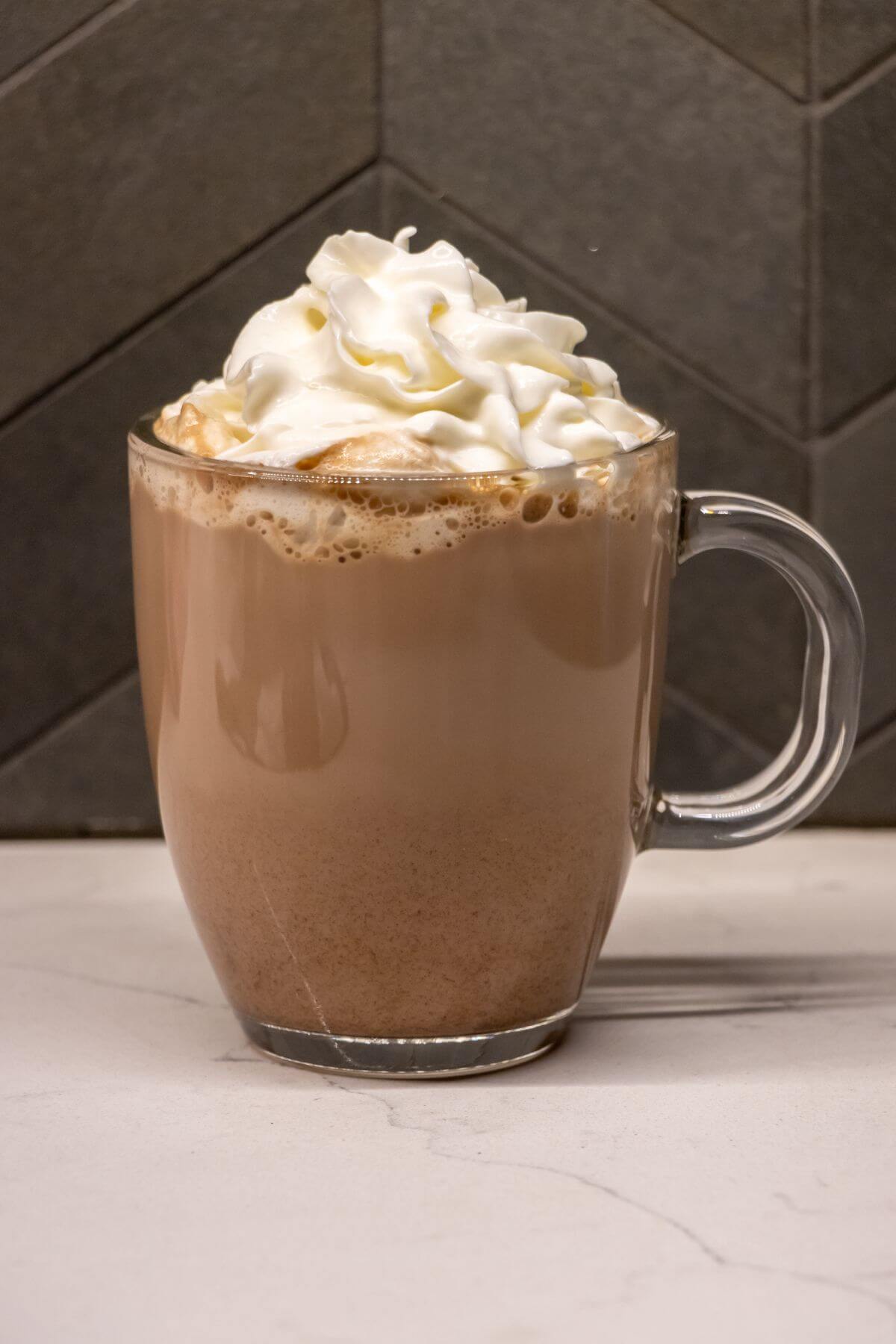 A glass mug filled with a frothy coffee drink, topped with a swirl of whipped cream, sits on a light-colored countertop in front of a tiled wall.