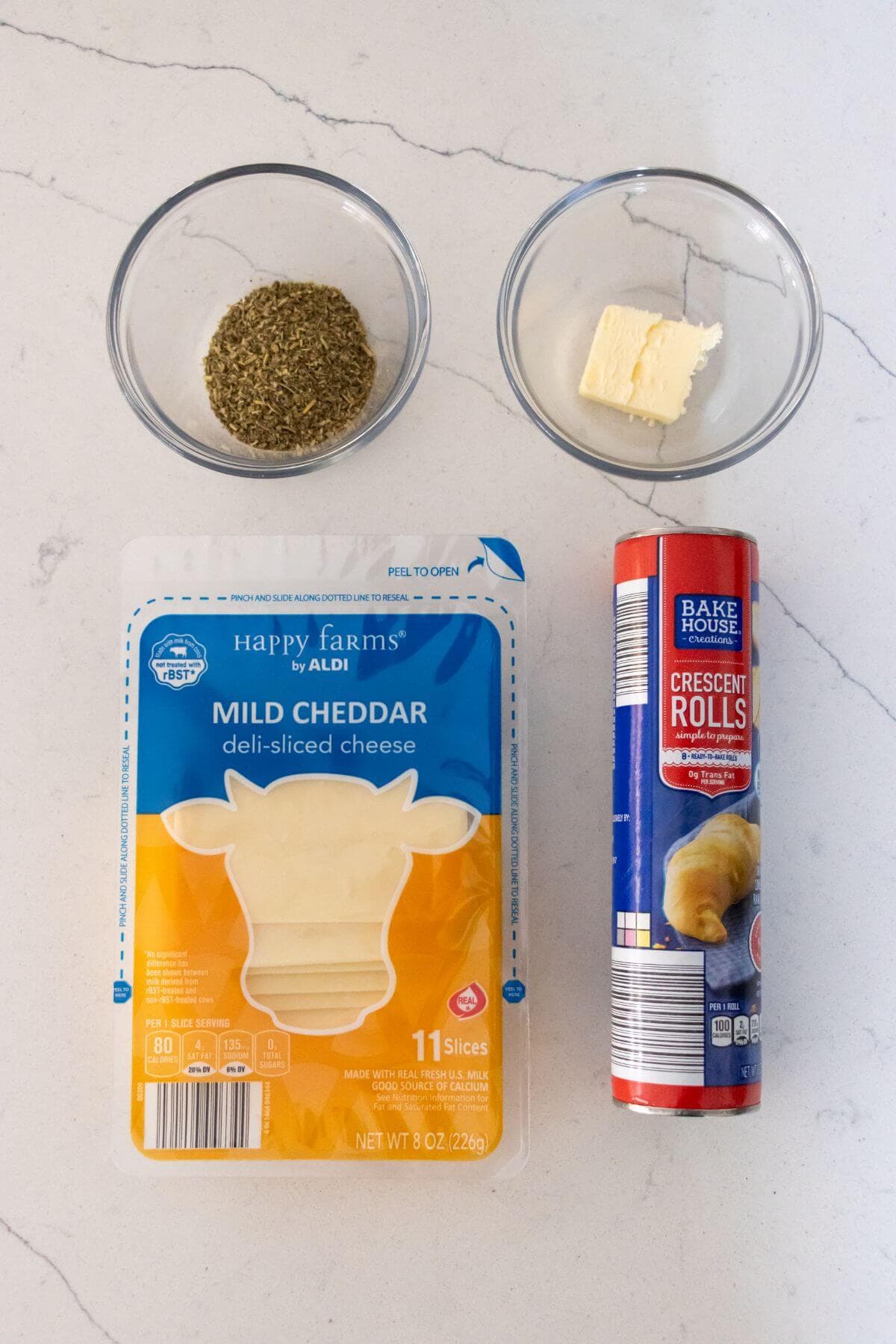 Package of mild cheddar cheese slices, a can of crescent rolls, a small bowl of dried herbs, and a small bowl of butter on a white surface.