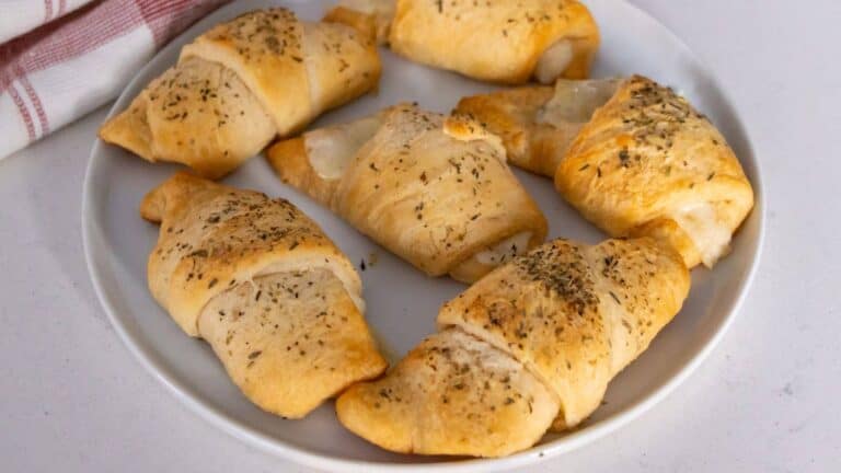 A white plate with six baked crescent rolls topped with dried herbs, placed on a white surface near a cloth napkin.
