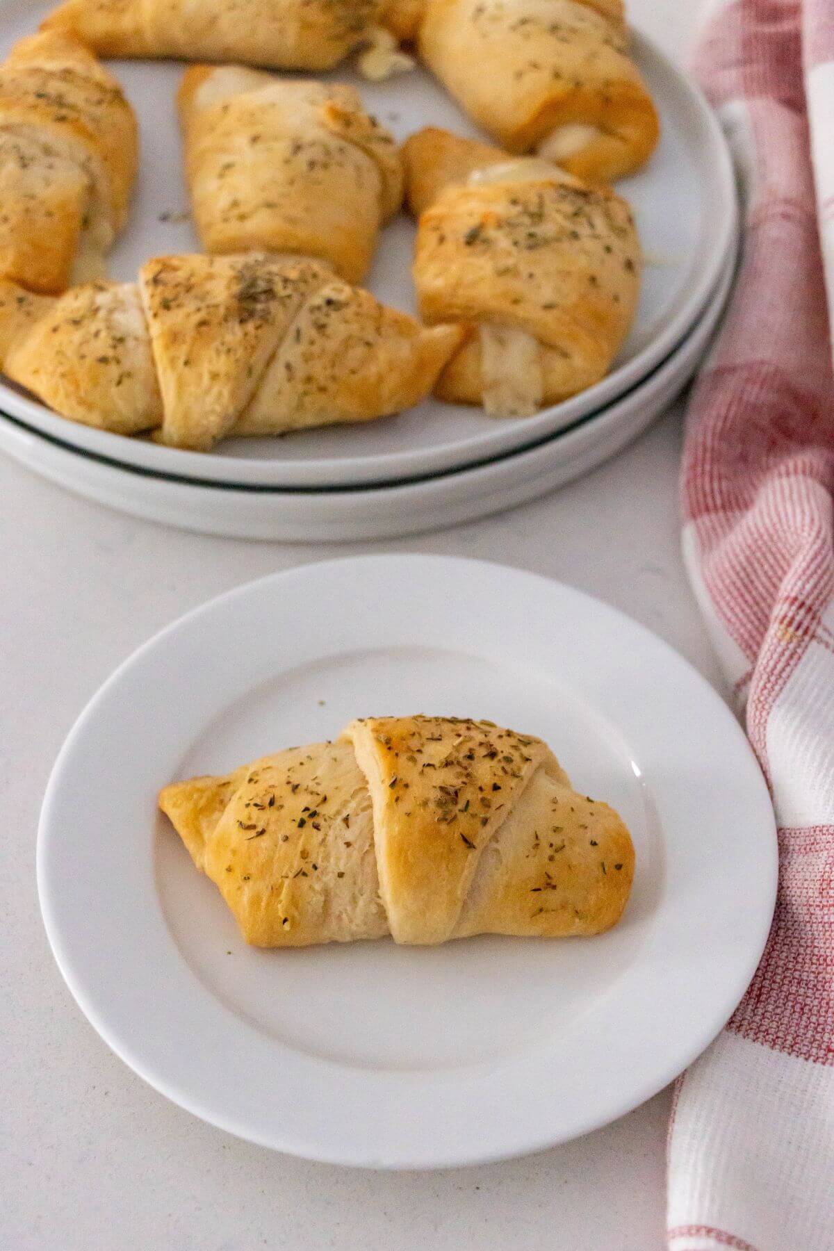 A plate with a single herb-topped crescent roll in the foreground and a larger plate with several crescent rolls in the background.