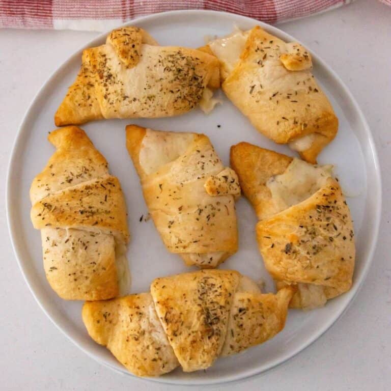 A white plate with six golden-brown crescent rolls topped with herbs, filled with melted cheese, on a light surface.