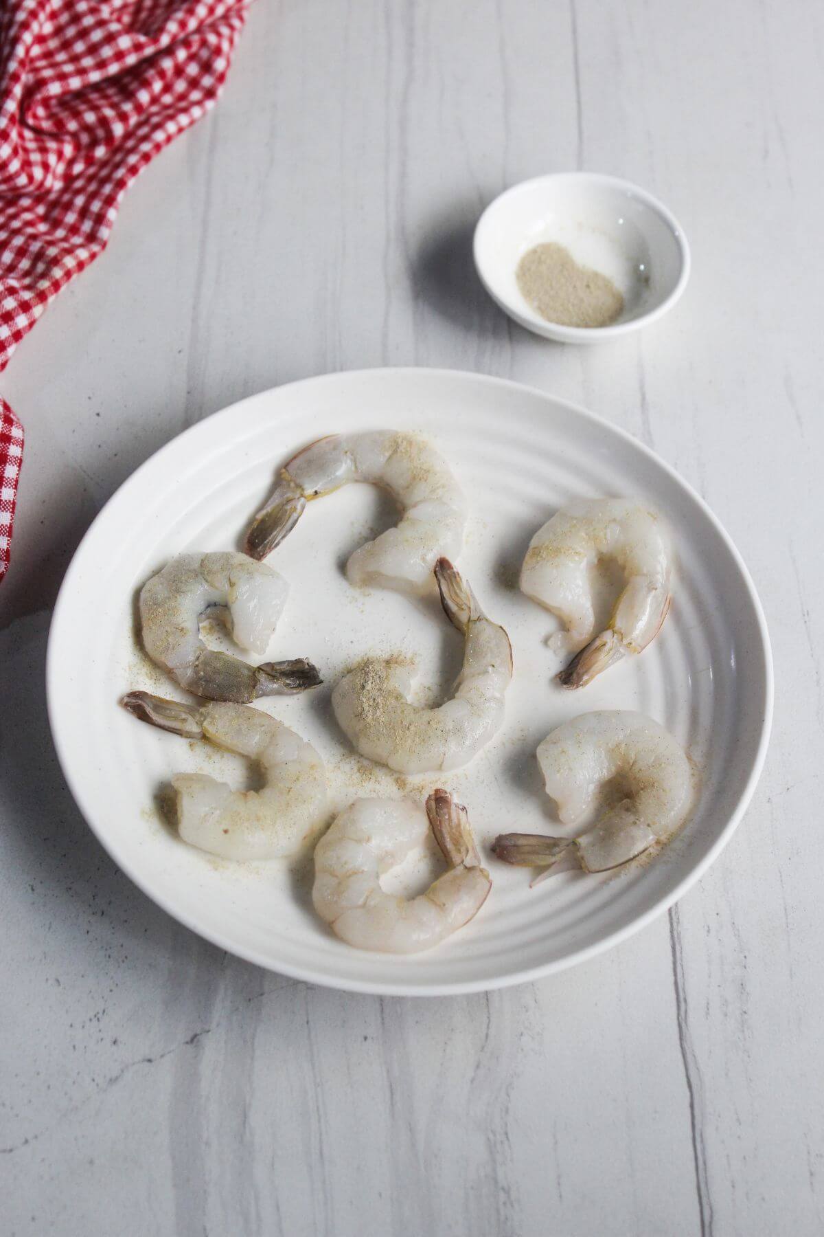 A white plate with six raw shrimp sprinkled with seasoning, set on a light surface. A small white bowl with more seasoning and a red checkered cloth are nearby.
