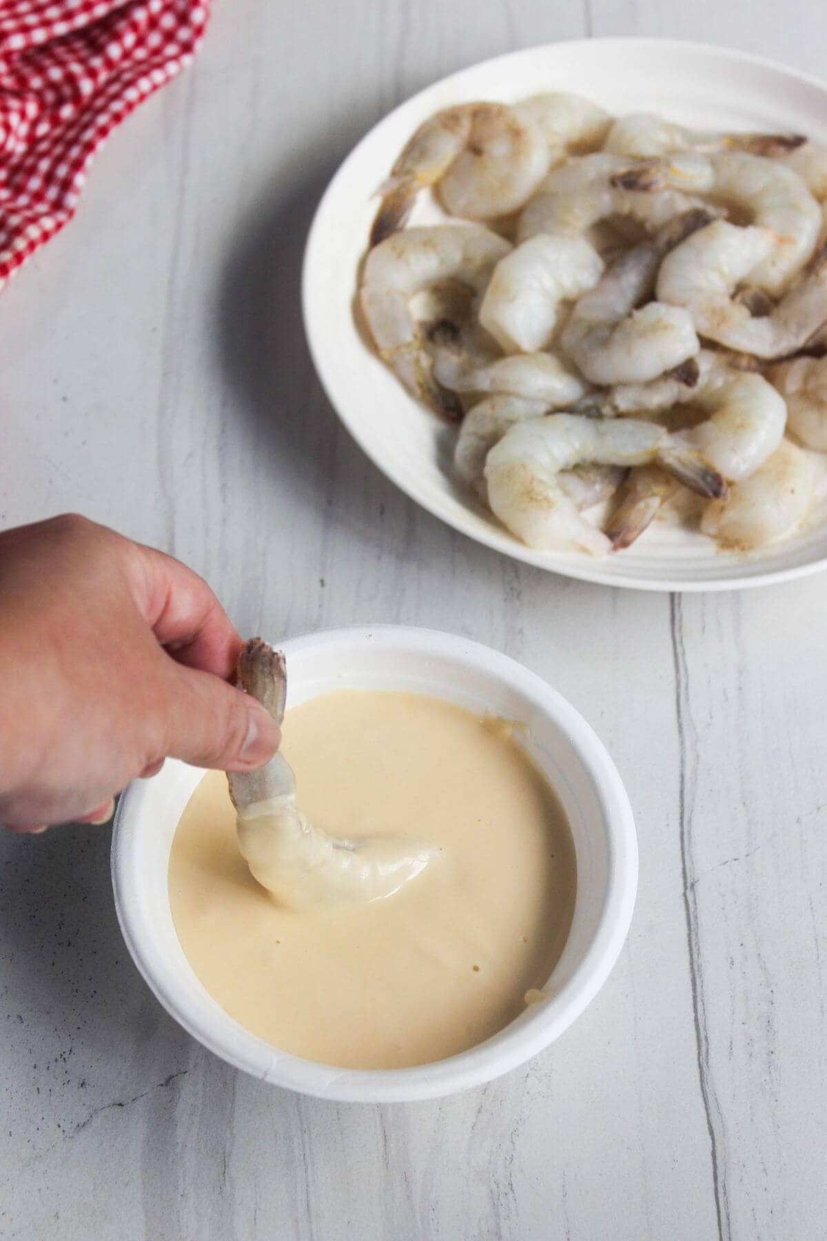 A hand dips a raw shrimp into a bowl of pale batter, with a plate of more raw shrimp in the background on a light countertop.